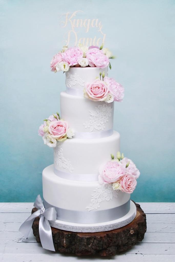 Czteropiętrowy tort weselny w stylu rustykalnym, dekorowany białym lukrem, koronkowymi zdobieniami, różowymi kwiatami i srebrną wstążką, umieszczony na drewnianym pniu z imionami Kinga i Daniel...
