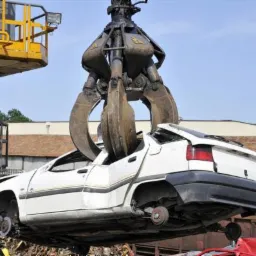 Uszkodzony, biały samochód osobowy podnoszony przez chwytak dźwigu na złomowisku w Poznaniu. Widoczne zgniecione elementy karoserii i brak kół.