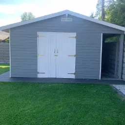 Szara, drewniana altana z białymi drzwiami dwuskrzydłowymi i złotymi zawiasami, widok z przodu na tle zielonego trawnika i częściowo widocznego ogrodzenia.