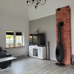 Jasne panele podłogowe w salonie z wysokim sufitem, kominkiem obudowanym czerwoną cegłą, białymi meblami RTV i czarnym żyrandolem