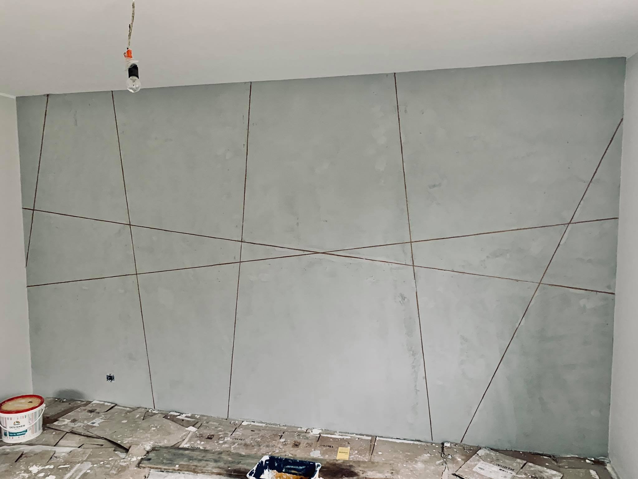 Szara ściana z dekoracyjnymi, geometrycznymi liniami w trakcie prac wykończeniowych; widoczne zabezpieczenia podłogi i puszka z farbą.