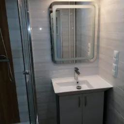 Nowoczesna łazienka po remoncie: kabina prysznicowa z drewnopodobnymi panelami, umywalka z szafką w kolorze gołębim, podświetlane lustro i płytki imitujące kamień na ścianach.