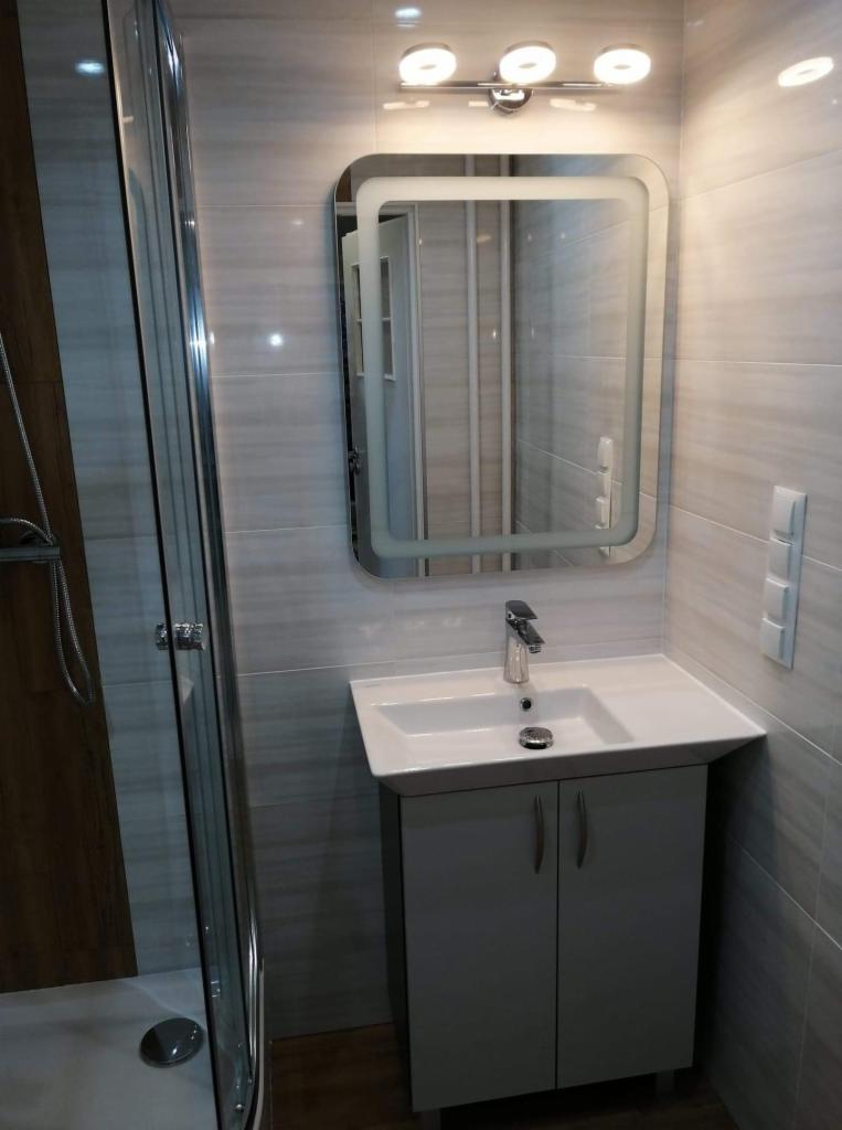 Nowoczesna łazienka po remoncie: kabina prysznicowa z drewnopodobnymi panelami, umywalka z szafką w kolorze gołębim, podświetlane lustro i płytki imitujące kamień na ścianach.