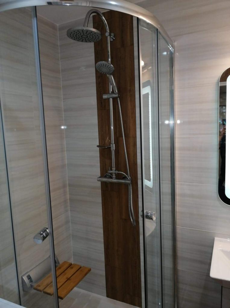 Nowoczesna kabina prysznicowa z deszczownicą, siedziskiem i panelem imitującym drewno, umieszczona w łazience wykończonej płytkami w odcieniach szarości.