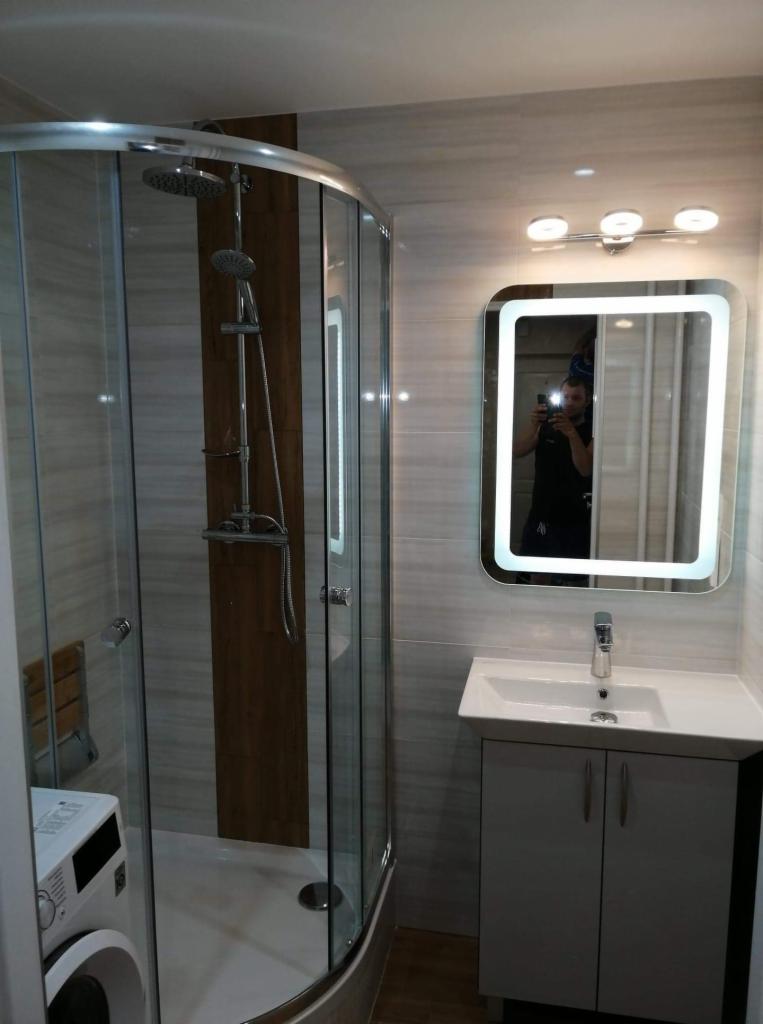 Wykończona łazienka z kabiną prysznicową z hydromasażem, umywalką z szafką i podświetlanym lustrem, widoczna pralka i refleksja fotografa w lustrze.