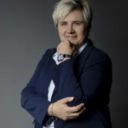 Portret uśmiechniętej kobiety w granatowej marynarce, opierającej podbródek na dłoni, z widocznym zegarkiem i złotą bransoletką na tle szarego studia.