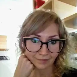 Portret nauczycielki w okularach na tle regału z książkami. Uśmiechnięta kobieta z blond włosami pozuje, opierając brodę na dłoni.