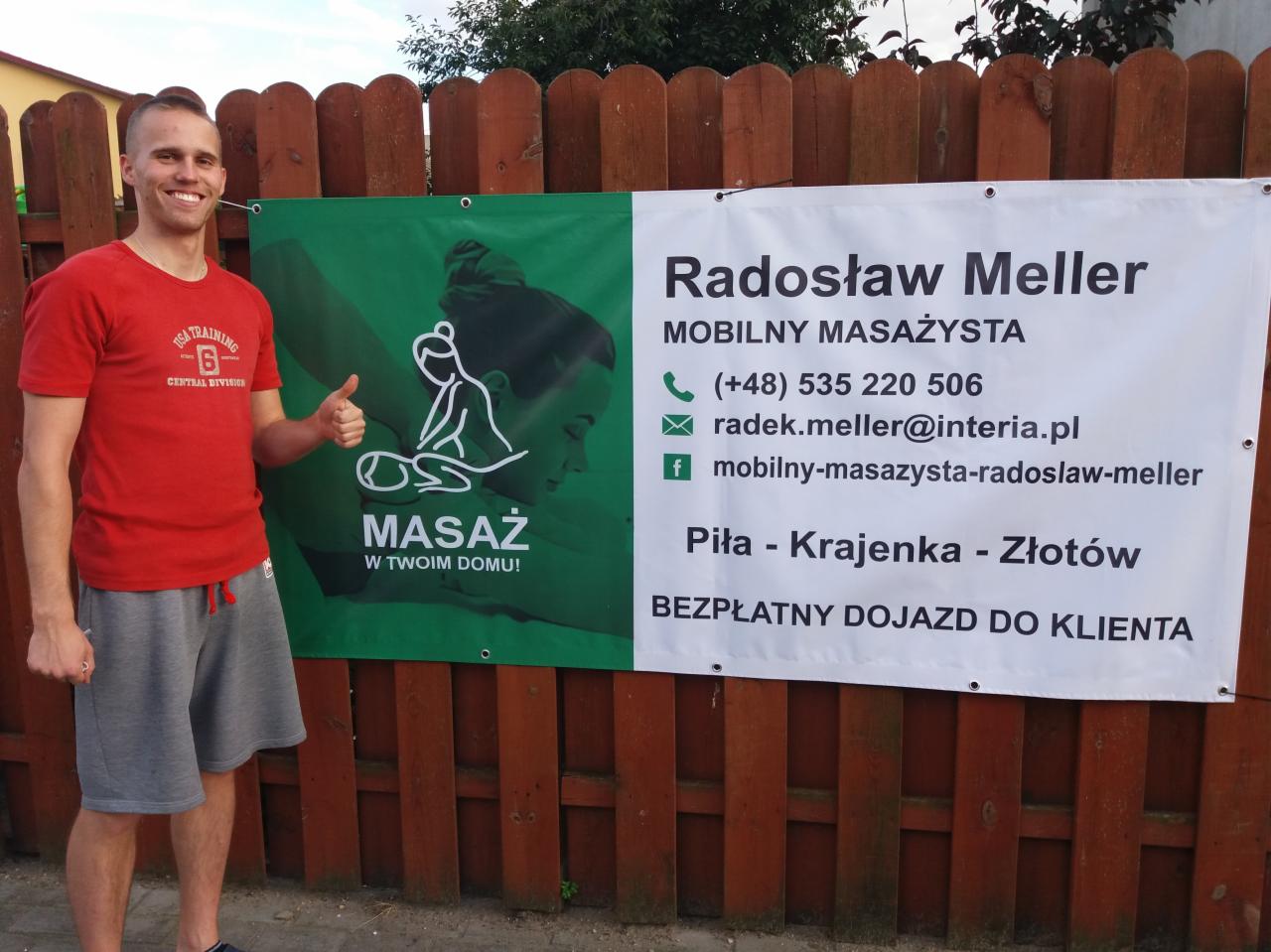 Uśmiechnięty mężczyzna w czerwonej koszulce i szarych spodenkach pozuje na tle drewnianego płotu z reklamą mobilnego masażysty Radosława Mellera, oferującego masaż w domu klienta na terenie Piły...