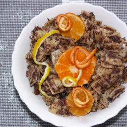 Kluski z makiem w białej misce, udekorowane skórką pomarańczy i cytryny, na błyszczącym, szarym tle.