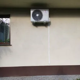 Zewnętrzna jednostka klimatyzacji marki Rotenso zamontowana na elewacji budynku, z widocznym kablem biegnącym w dół ściany, obok okna z firanką i roślinami.