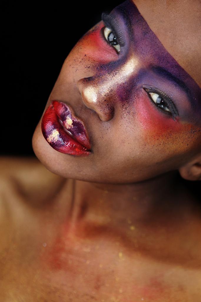 Portret kobiety z artystycznym makijażem w odcieniach fioletu, czerwieni i złota, z bliska, na czarnym tle. Usta pomalowane intensywną, błyszczącą szminką.