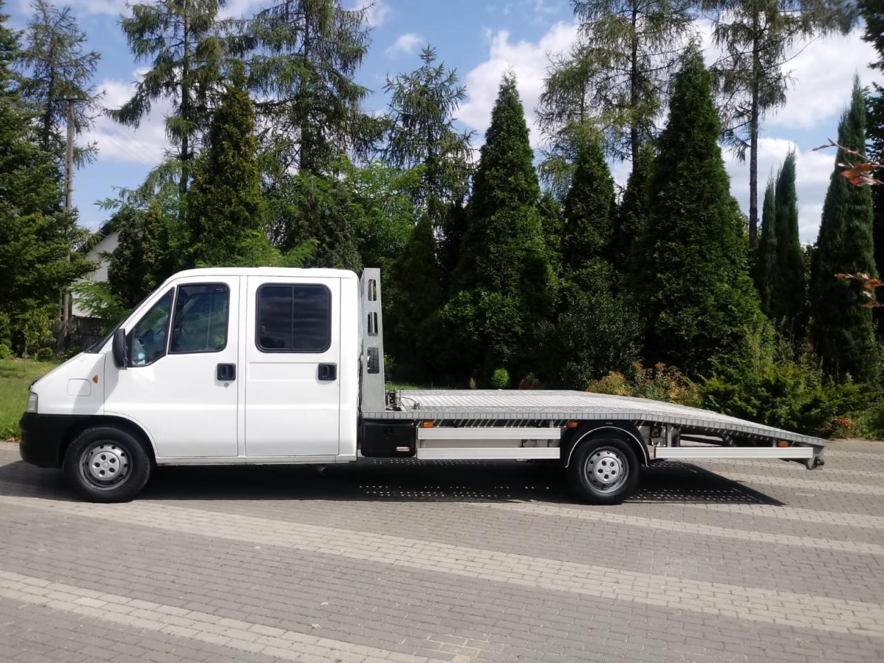 Biała laweta z kabiną załogową, zaparkowana na kostce brukowej na tle zielonych drzew i błękitnego nieba.