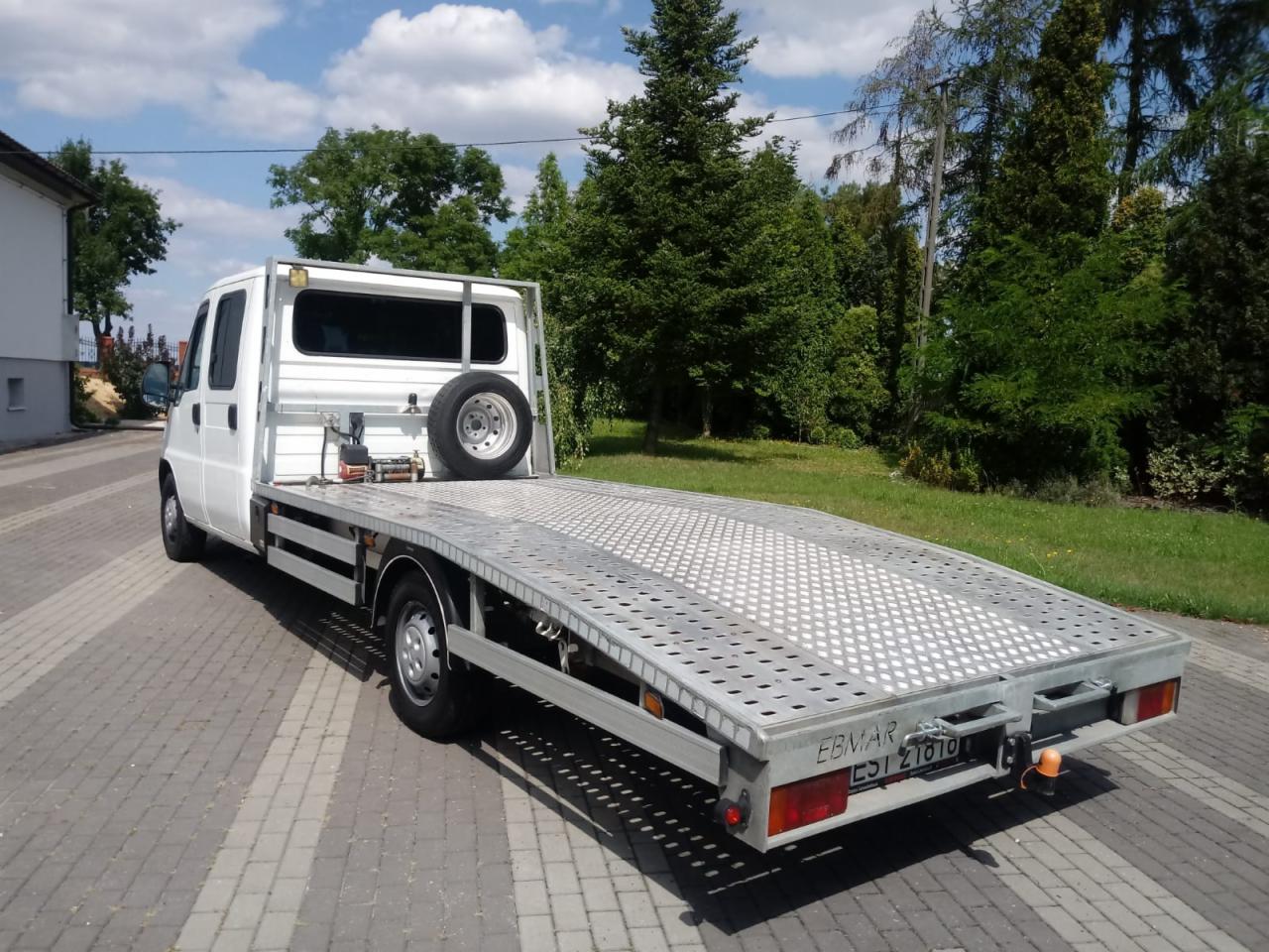 Biała laweta z metalową platformą do transportu pojazdów, zaparkowana na brukowanej powierzchni w słoneczny dzień.