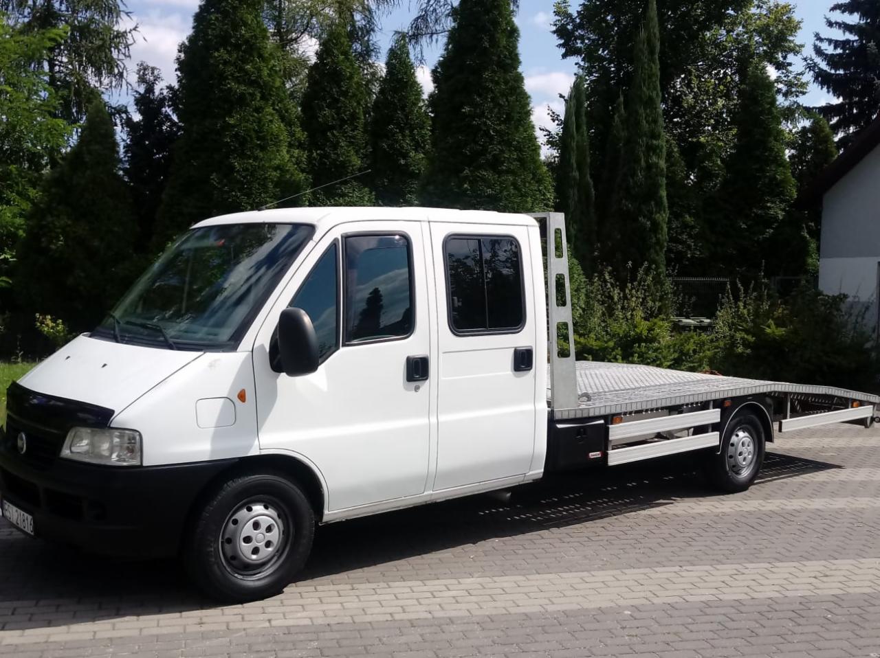 Biała laweta na tle zielonych drzew, zaparkowana na brukowanej nawierzchni.