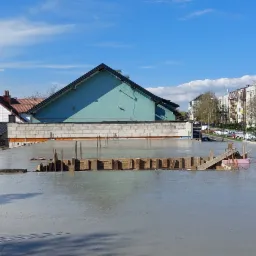 Świeżo wylana, gładka powierzchnia betonowa z wystającymi prętami zbrojeniowymi i drewnianym szalunkiem w tle, widok na zabudowę miejską w słoneczny dzień.