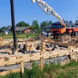 Wykop pod fundamenty z drewnianym szalunkiem, beton wylewany z pompy, w tle domy jednorodzinne i błękitne niebo.