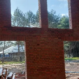 Surowa ściana z czerwonej cegły z trzema otworami okiennymi i otworem drzwiowym, widok na las i altanę w tle, drewniana konstrukcja dachu w górnej części kadru.