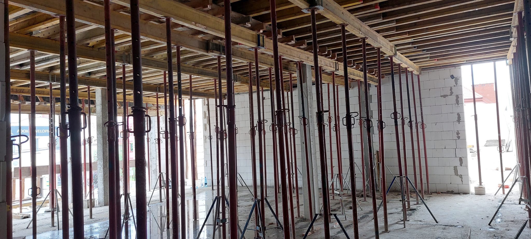Wnętrze w trakcie budowy z widocznymi podporami stropowymi, drewnianymi belkami i fragmentem murowanej ściany z bloczków betonu komórkowego.