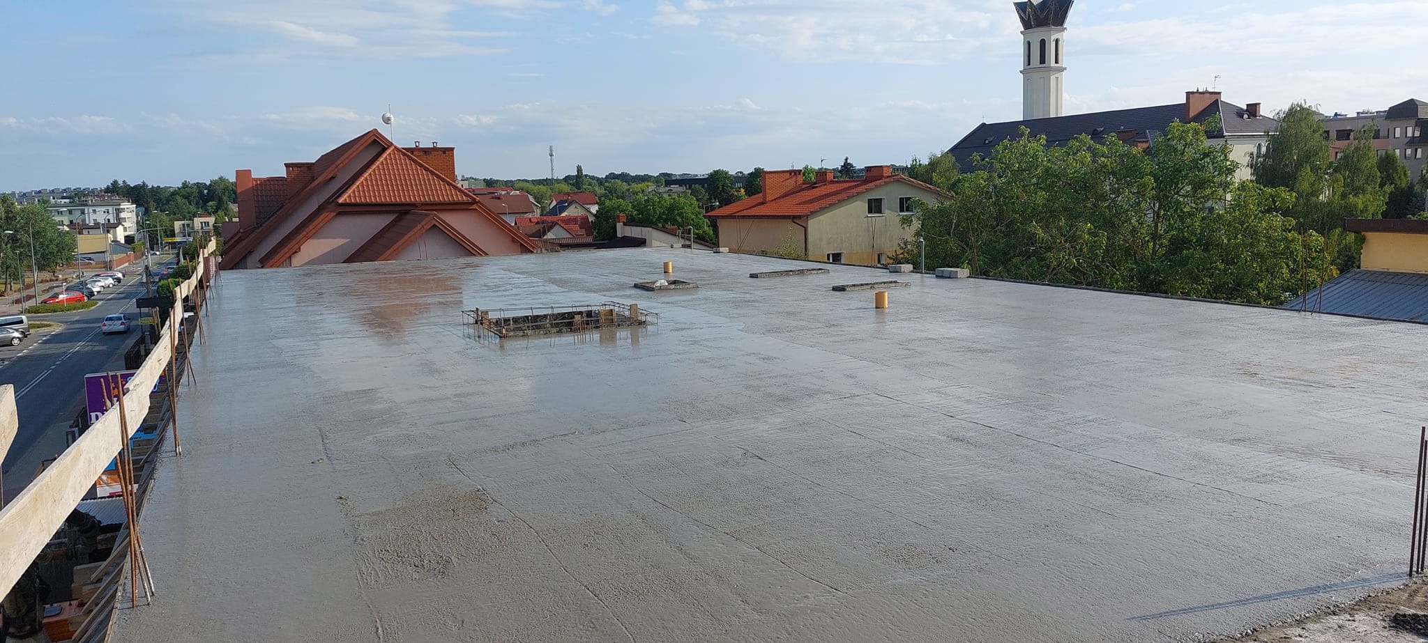 Świeżo wylana, mokra wylewka na płaskim dachu z widoczną zbrojoną ramą włazu i żółtymi ogranicznikami, w tle zabudowa miejska i kościół.