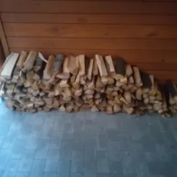 Ułożony stos pociętego drewna opałowego na szarym brukowanym podłożu, przylegający do drewnianej ściany z desek.