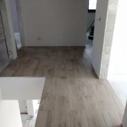 Ułożona podłoga z płytek imitujących drewno w jasnym odcieniu, widok z g&oacute;ry na fragment pomieszczenia w trakcie remontu, z widocznymi surowymi ścianami i fragmentami konstrukcji z płyt...