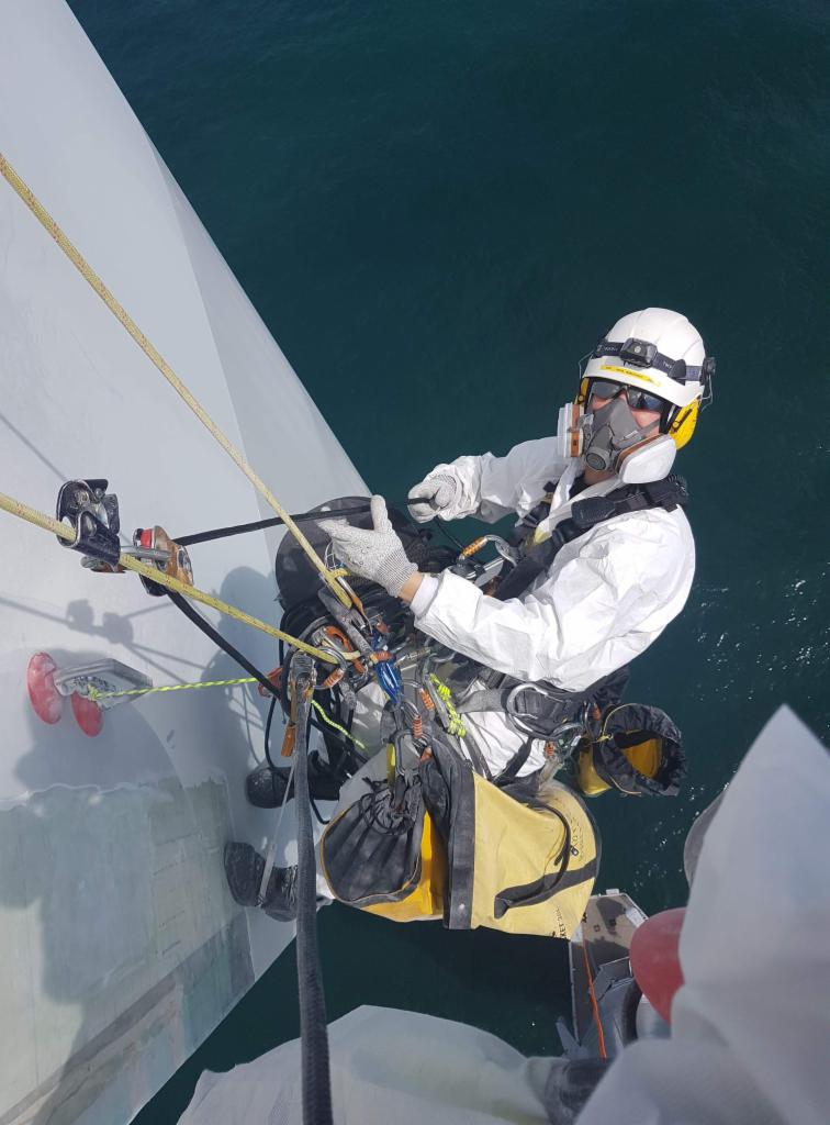 Pracownik w kombinezonie, kasku i masce, zabezpieczony linami, wykonuje prace na wysokości na tle morza. Widoczne narzędzia i sprzęt specjalistyczny.