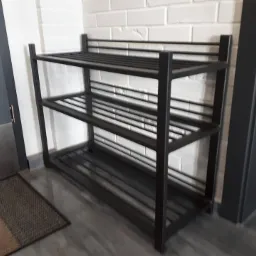 Metalowy regał na buty z trzema półkami, ustawiony przy ścianie z białej cegły i obok ciemnoszarych drzwi.