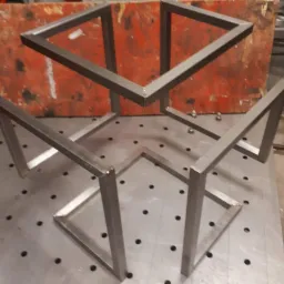 Metalowa konstrukcja przestrzenna z profili kwadratowych, spawana, surowy stan, widoczne spawy, tło z pomarańczową płytą i metalowy stół spawalniczy z otworami.