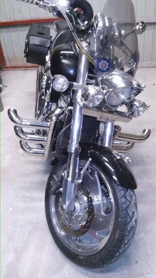 Częściowo widoczny motocykl typu cruiser z chromowanymi elementami, w tym reflektory, orurowanie ochronne i przedni błotnik, z dodatkową torbą boczną.