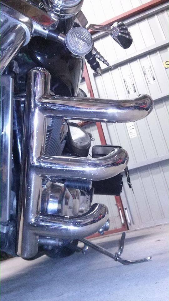 Chromowana rura ochronna motocykla zamontowana na czarnej maszynie, widoczna w warsztacie z metalowymi ścianami.