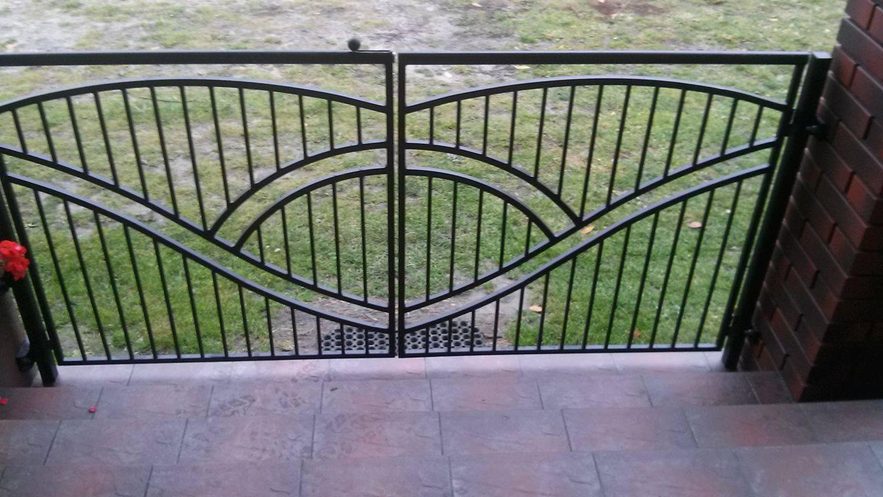 Metalowa brama dwuskrzydłowa z dekoracyjnymi łukami, zamontowana na podeście z płytek, widok na trawnik w tle.