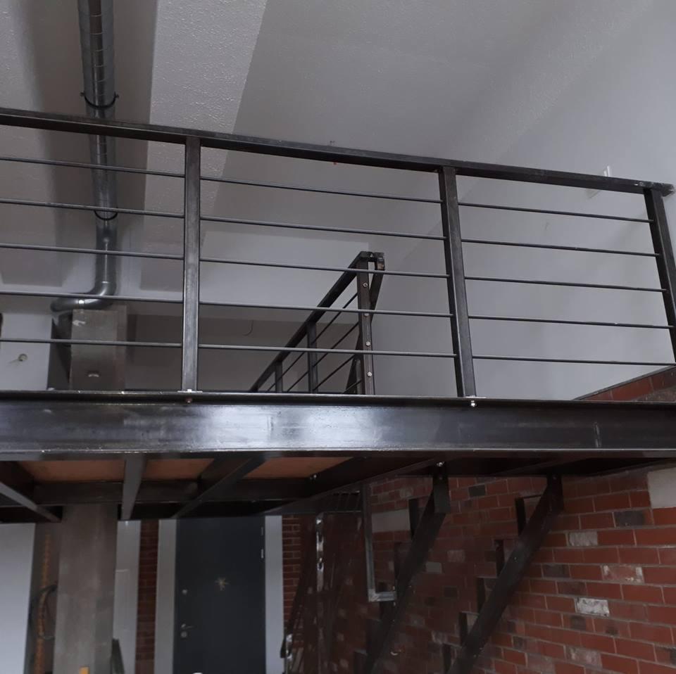 Metalowa konstrukcja antresoli z balustradą z poziomych prętów, widoczne elementy mocowania do ściany z cegły, szara kolorystyka, surowy industrialny styl.