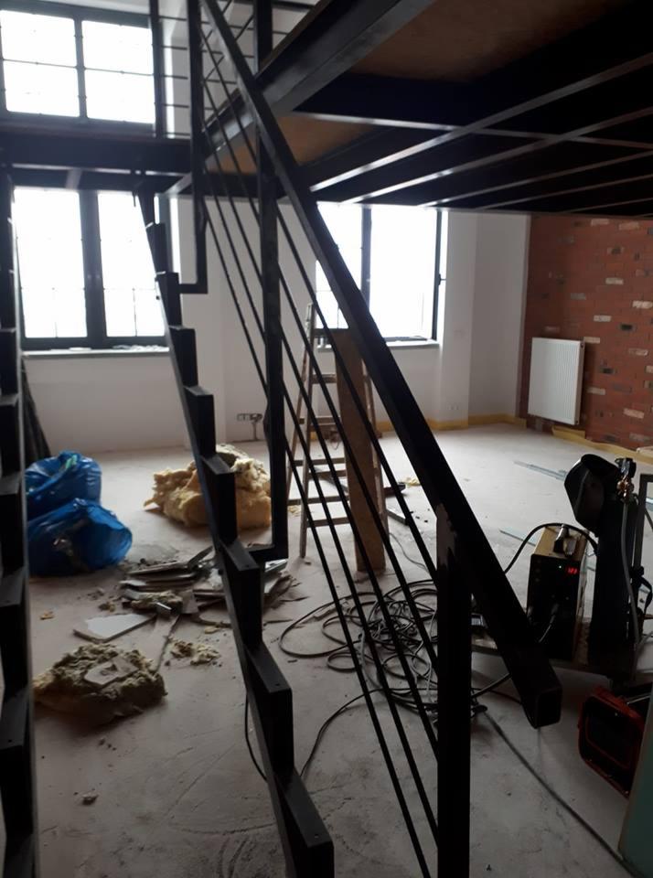 Metalowe schody z balustradą w stylu loft, widoczne ślady prac spawalniczych, sprzęt spawalniczy, surowy stan wykończenia wnętrza z ceglaną ścianą w tle.