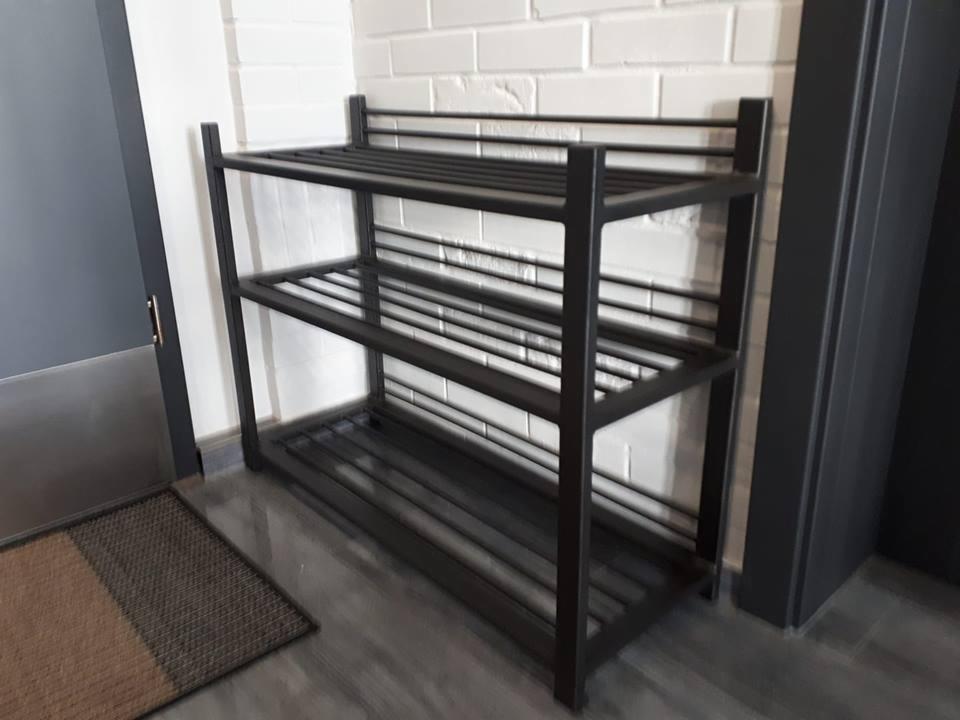 Metalowy regał na buty z trzema półkami, ustawiony przy ścianie z białej cegły i obok ciemnoszarych drzwi.