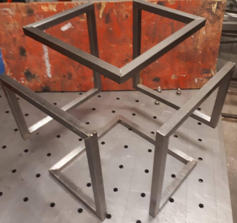 Metalowa konstrukcja przestrzenna z profili kwadratowych, spawana, surowy stan, widoczne spawy, tło z pomarańczową płytą i metalowy stół spawalniczy z otworami.