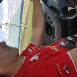 Mężczyzna w czerwonej koszulce siedzi w samochodzie, widok przez otwarte okno na pole i słup energetyczny. Nietypowy kąt ujęcia.