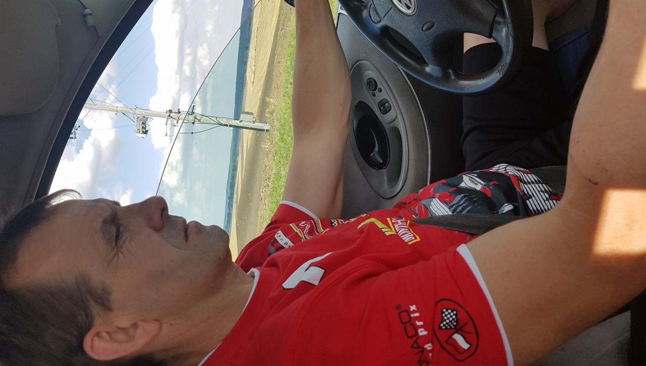 Mężczyzna w czerwonej koszulce siedzi w samochodzie, widok przez otwarte okno na pole i słup energetyczny. Nietypowy kąt ujęcia.