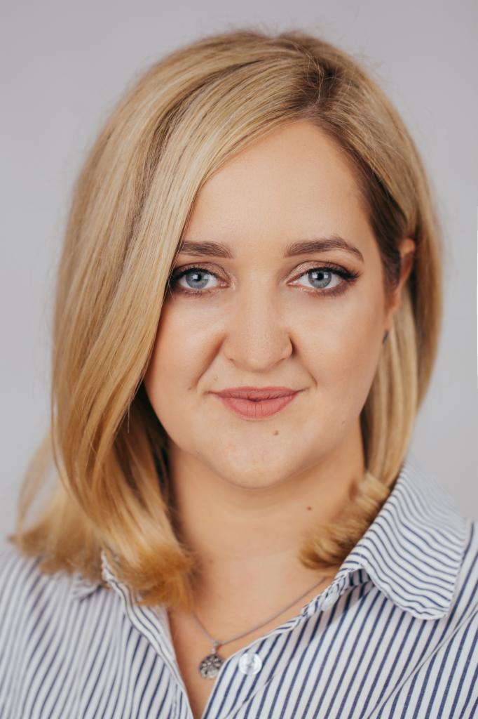 Portret kobiety z blond włosami, niebieskimi oczami i delikatnym uśmiechem, ubrana w koszulę w paski, na jasnoszarym tle.