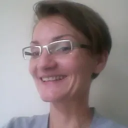 Uśmiechnięta kobieta w okularach z metalową oprawką, krótko ostrzyżona, w szarej bluzce, na jasnym tle.