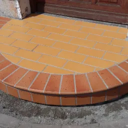 Półokrągłe schody wejściowe wyłożone pomarańczową terakotą i ceglaną obwódką w Dzierżoniowie, widoczne ślady prac wykończeniowych wokół.