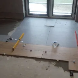 Układanie drewnopodobnych płytek podłogowych w trakcie prac wykończeniowych, widoczne poziomice, krzyżyki dystansowe i zapasowe płytki przy oknie.