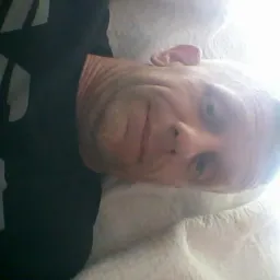 Portret mężczyzny leżącego na białej pościeli, z uśmiechem i spojrzeniem w obiektyw, w czarnej koszulce. Zdjęcie obrócone.