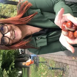 Uśmiechnięta kobieta w okularach i zielonym płaszczu trzyma w dłoniach kilka kasztanów na tle parku z ławkami.