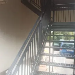 Zewnętrzne schody metalowe z ażurowymi stopniami i prostymi balustradami, prowadzące na wyższą kondygnację budynku, z zaparkowanym niebieskim samochodem w tle.