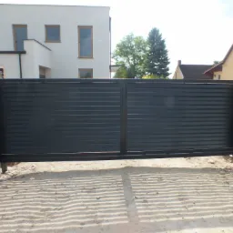 Czarna, pozioma brama żaluzjowa zamontowana między szarymi słupkami z betonowych bloczków, na tle jasnego domu i piaszczystego podłoża.