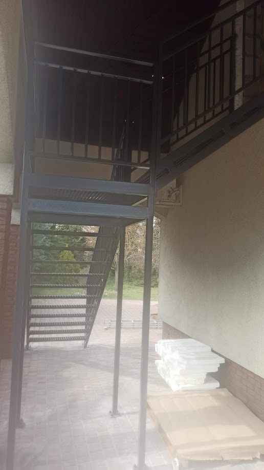 Zewnętrzne schody metalowe z czarną balustradą prowadzące na piętro budynku, widoczne z poziomu brukowanego podwórza, z zapasem materiałów budowlanych w pobliżu.