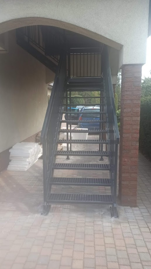 Zewnętrzne schody metalowe z kratownicowymi stopniami, prowadzące na górę, z widoczną konstrukcją podtrzymującą i fragmentem ceglanej kolumny.