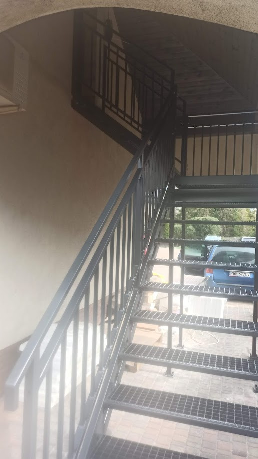 Zewnętrzne schody metalowe z ażurowymi stopniami i prostymi balustradami, prowadzące na wyższą kondygnację budynku, z zaparkowanym niebieskim samochodem w tle.