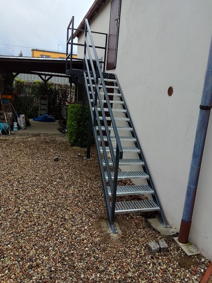 Zewnętrzne schody metalowe z kratowymi stopniami, prowadzące do drzwi na piętrze budynku, widoczne na tle żwirowego podłoża.