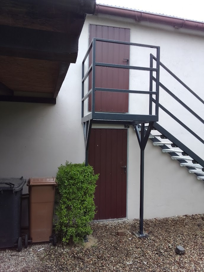 Zewnętrzne schody metalowe z balustradą prowadzące do drzwi na piętrze, widoczne kosze na śmieci i mały krzew u podstawy.